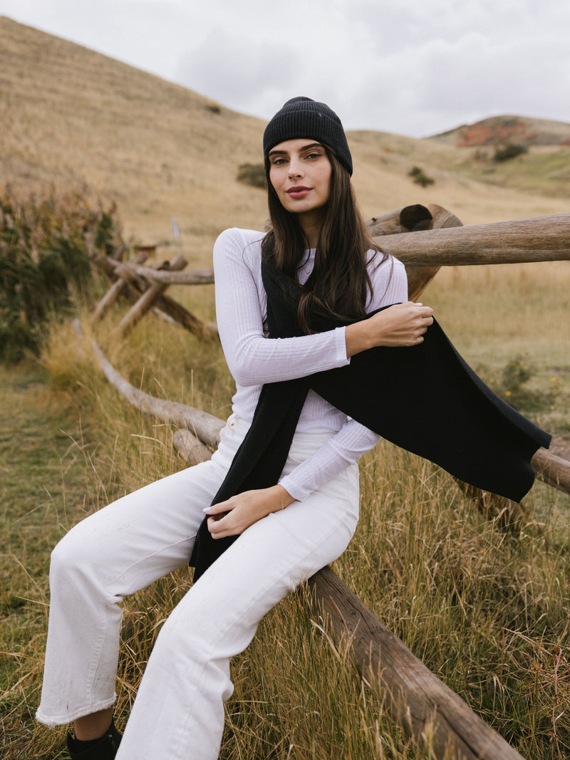 Woman wearing a black knit beanie and scarf sits on a wooden fence in a field