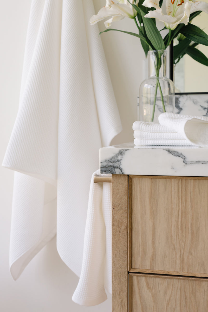 White Waffle Hand Towel hanging from a gold towel bar. The photo was taken at a white marble bathroom sink. A Waffle Bath Towel is seen hanging on the wall in the background.